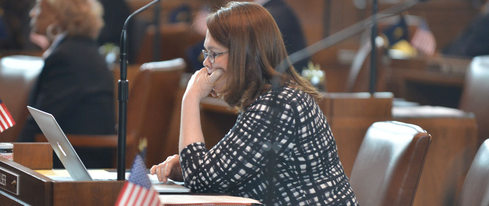 A white woman is viewed from the side.  She is sitting at a desk with her chin resting on her hand.  She is wearing a black checked shirt and glasses.