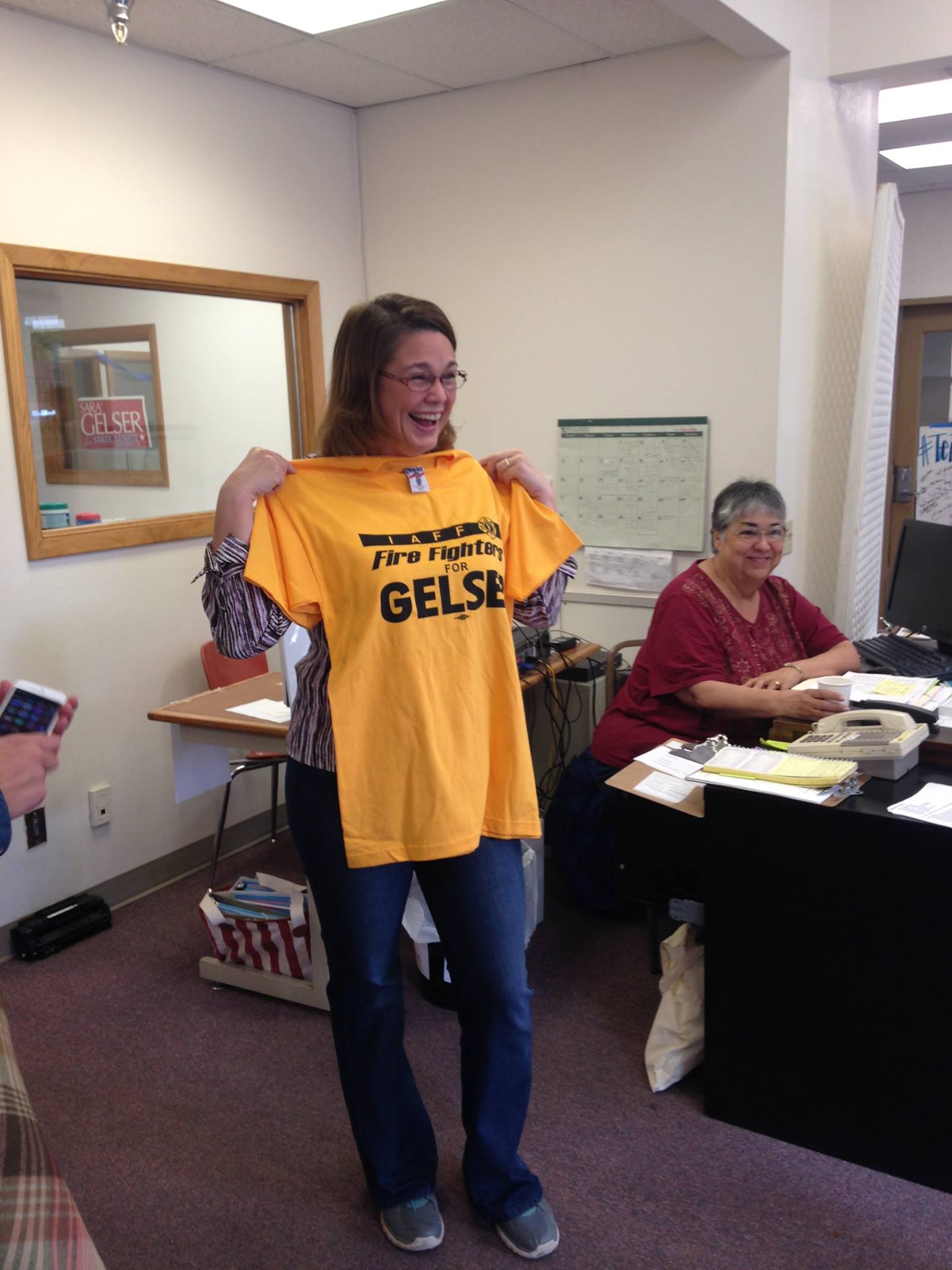 A white woman with dark hair is wearing glasses and blue jeans. She is smiling and holding up a shirt that says Firefighters for Gelser. In the background, a smiling woman at a desk looks on.