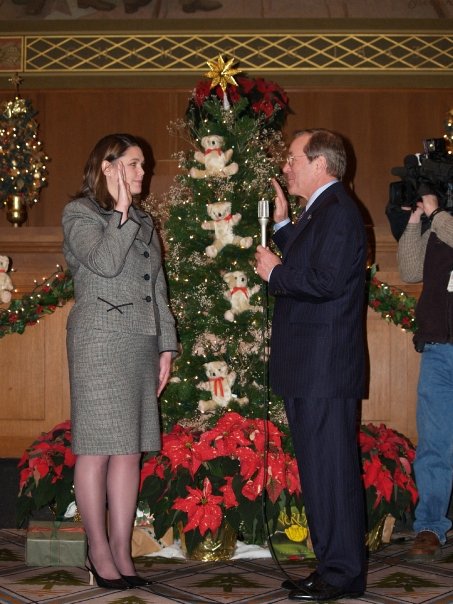 Two people stand in front of a decorated Christmas tree and a mound of poinsettias. On the right is a white man with a dark suit. On the left is a white woman with dark hair. She is wearing a checkered suit and black heels. Her right hand is raised to take an oath.
